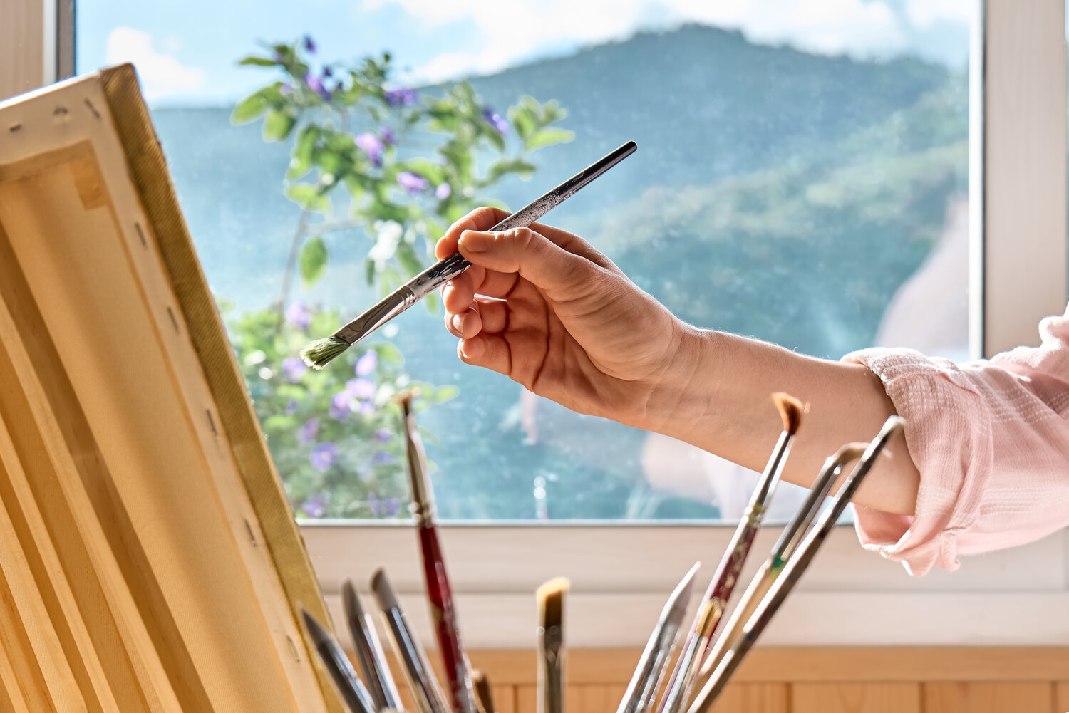 Female hand holding a paintbrush near canvas on easel near the window in the studio.