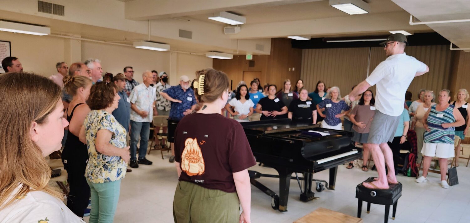 Photo of Ben with the choir during their first practice of the season, as he leads a vocal warmup and breathing exercises. 2023.