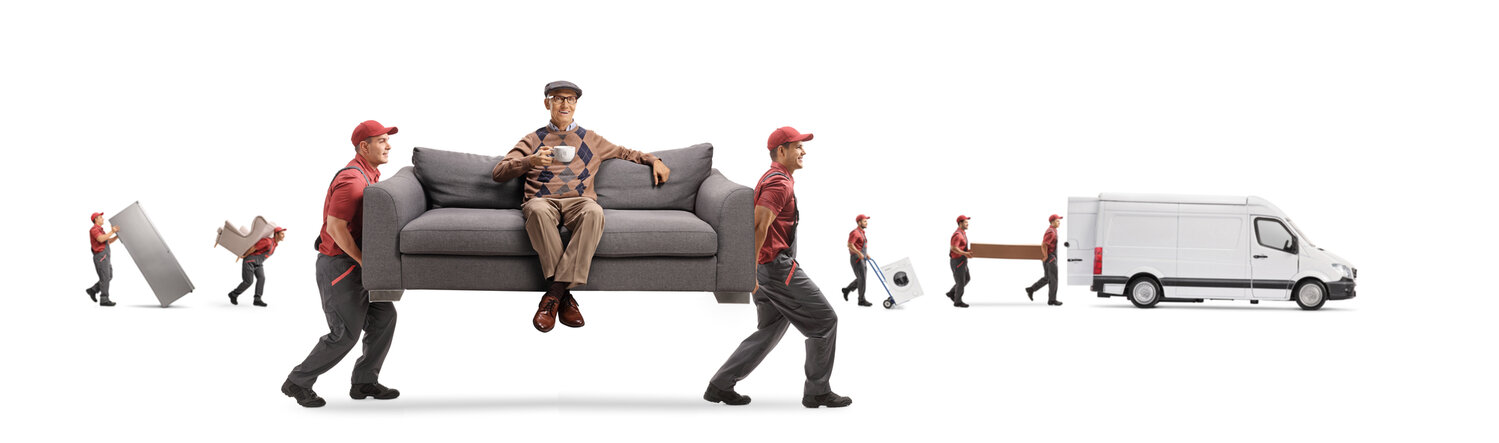 Movers moving a couch which an an elderly man is sitting on, while he holds a cup of tea and smiles